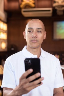 Beyaz polo tişörtlü kel Latin bir adamın portresi, şık bir Çin tarzı kahve dükkanında oturuyor, ev ortamında kendine güveni tam görünüyor..