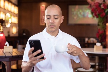 Beyaz polo tişörtlü kel Latin bir adamın portresi, şık bir Çin tarzı kahve dükkanında oturuyor, ev ortamında kendine güveni tam görünüyor..