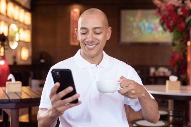 Beyaz polo tişörtlü kel Latin bir adamın portresi, şık bir Çin tarzı kahve dükkanında oturuyor, ev ortamında kendine güveni tam görünüyor..