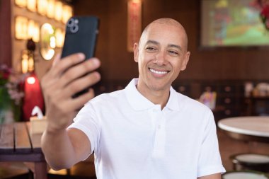 Beyaz polo tişörtlü kel Latin bir adamın portresi, şık bir Çin tarzı kahve dükkanında oturuyor, ev ortamında kendine güveni tam görünüyor..