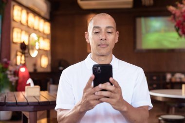 Beyaz polo tişörtlü kel Latin bir adamın portresi, şık bir Çin tarzı kahve dükkanında oturuyor, ev ortamında kendine güveni tam görünüyor..