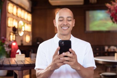 Beyaz polo tişörtlü kel Latin bir adamın portresi, şık bir Çin tarzı kahve dükkanında oturuyor, ev ortamında kendine güveni tam görünüyor..