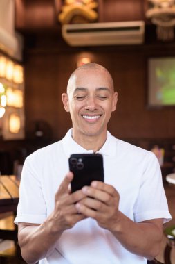 Beyaz polo tişörtlü kel Latin bir adamın portresi, şık bir Çin tarzı kahve dükkanında oturuyor, ev ortamında kendine güveni tam görünüyor..