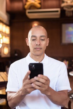 Beyaz polo tişörtlü kel Latin bir adamın portresi, şık bir Çin tarzı kahve dükkanında oturuyor, ev ortamında kendine güveni tam görünüyor..