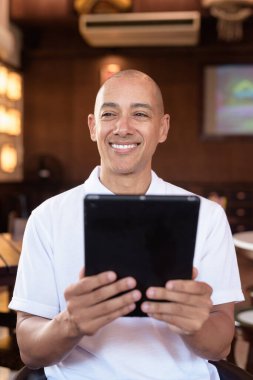 Beyaz polo tişörtlü kel Latin bir adamın portresi, şık bir Çin tarzı kahve dükkanında oturuyor, ev ortamında kendine güveni tam görünüyor..