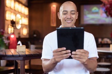 Beyaz polo tişörtlü kel Latin bir adamın portresi, şık bir Çin tarzı kahve dükkanında oturuyor, ev ortamında kendine güveni tam görünüyor..