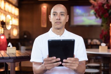 Beyaz polo tişörtlü kel Latin bir adamın portresi, şık bir Çin tarzı kahve dükkanında oturuyor, ev ortamında kendine güveni tam görünüyor..