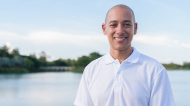 Sakin bir gölün kenarında duran beyaz polo tişörtlü şık kel bir adam. Huzurlu deniz manzaralı doğal bir açık hava portresi içinde kendinden emin ve rahat bir ifade..