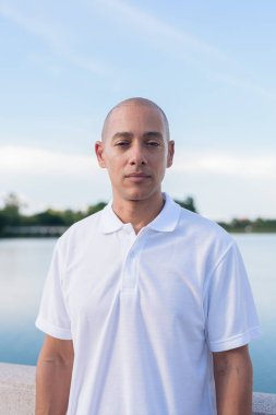 Sakin bir gölün kenarında duran beyaz polo tişörtlü şık kel bir adam. Huzurlu deniz manzaralı doğal bir açık hava portresi içinde kendinden emin ve rahat bir ifade..