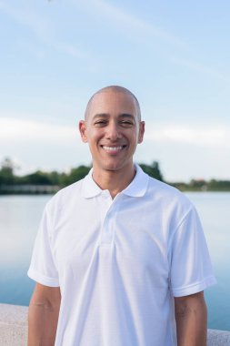 Sakin bir gölün kenarında duran beyaz polo tişörtlü şık kel bir adam. Huzurlu deniz manzaralı doğal bir açık hava portresi içinde kendinden emin ve rahat bir ifade..
