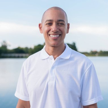 Sakin bir gölün kenarında duran beyaz polo tişörtlü şık kel bir adam. Huzurlu deniz manzaralı doğal bir açık hava portresi içinde kendinden emin ve rahat bir ifade..