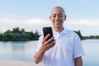 Beyaz polo tişörtlü şık kel adam akıllı telefon kullanarak sakin bir gölün kenarında dikiliyor. Huzurlu deniz manzaralı doğal bir açık hava portresi içinde kendinden emin ve rahat bir ifade..