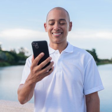 Beyaz polo tişörtlü şık kel adam akıllı telefon kullanarak sakin bir gölün kenarında dikiliyor. Huzurlu deniz manzaralı doğal bir açık hava portresi içinde kendinden emin ve rahat bir ifade..