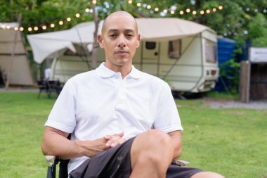 Beyaz polo tişörtlü şık kel bir adam kamp alanında karavanın yanında dinleniyor. Açık hava seyahati ve kamp yaşam tarzı konsepti çimenli çevre ve doğal ışık.