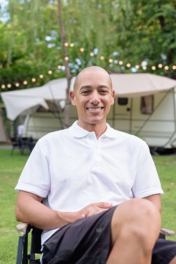 Beyaz polo tişörtlü şık kel bir adam kamp alanında karavanın yanında dinleniyor. Açık hava seyahati ve kamp yaşam tarzı konsepti çimenli çevre ve doğal ışık.