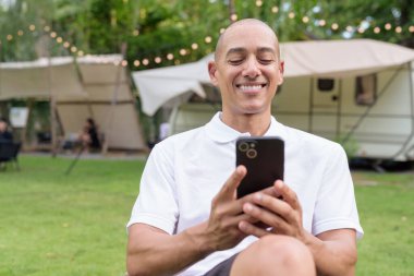 Beyaz polo tişörtlü şık kel bir adam kamp alanında karavanın yanında dinleniyor. Açık hava seyahati ve kamp yaşam tarzı konsepti çimenli çevre ve doğal ışık.