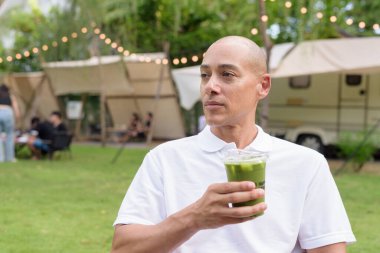 Beyaz polo tişörtlü şık kel bir adam kamp alanında karavanın yanında dinleniyor. Açık hava seyahati ve kamp yaşam tarzı konsepti çimenli çevre ve doğal ışık.