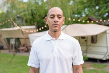 Beyaz polo tişörtlü şık kel bir adam kamp alanında karavanın yanında dinleniyor. Açık hava seyahati ve kamp yaşam tarzı konsepti çimenli çevre ve doğal ışık.