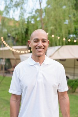 Beyaz polo tişörtlü şık kel bir adam kamp alanında karavanın yanında dinleniyor. Açık hava seyahati ve kamp yaşam tarzı konsepti çimenli çevre ve doğal ışık.