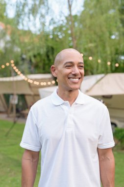 Beyaz polo tişörtlü şık kel bir adam kamp alanında karavanın yanında dinleniyor. Açık hava seyahati ve kamp yaşam tarzı konsepti çimenli çevre ve doğal ışık.