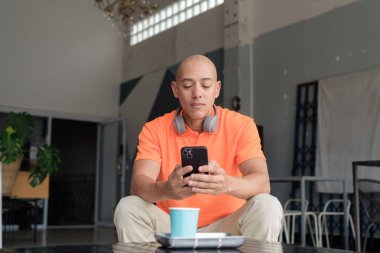 Kafe masasında kahve fincanı ile turuncu polo tişörtlü, kulaklık takan ve akıllı telefon kullanan orta yaşlı yakışıklı kel bir adam. Teknoloji ve yaşam tarzı kavramı.