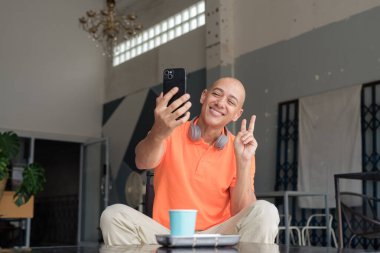 Kafe masasında kahve fincanı ile turuncu polo tişörtlü, kulaklık takan ve akıllı telefon kullanarak selfie çeken orta yaşlı yakışıklı kel adam. Teknoloji ve yaşam tarzı kavramı.