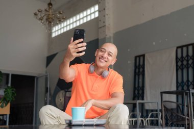 Kafe masasında kahve fincanı ile turuncu polo tişörtlü, kulaklık takan ve akıllı telefon kullanan orta yaşlı yakışıklı kel bir adam. Teknoloji ve yaşam tarzı kavramı.