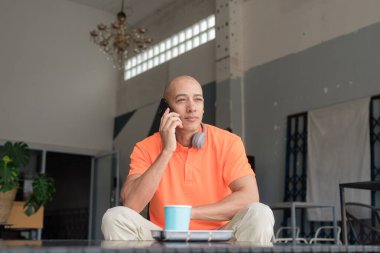 Kafe masasında kahve fincanı ile turuncu polo tişörtlü, kulaklık takan ve akıllı telefon kullanan orta yaşlı yakışıklı kel bir adam. Teknoloji ve yaşam tarzı kavramı.