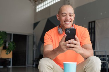 Kafe masasında kahve fincanı ile turuncu polo tişörtlü, kulaklık takan ve akıllı telefon kullanan orta yaşlı yakışıklı kel bir adam. Teknoloji ve yaşam tarzı kavramı.