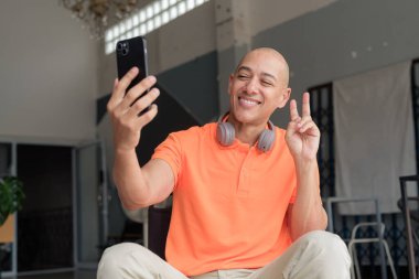 Kel İspanyol adam, portakal rengi polo tişört giyerek kafede otururken bir yandan da selfie çekiyor ve barış işareti yapıyor.