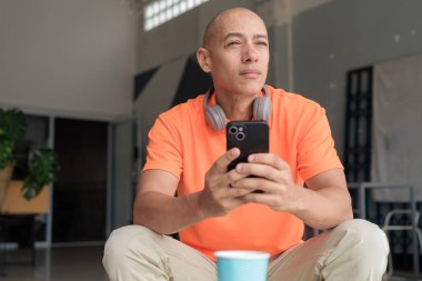 Kafe masasında kahve fincanı ile turuncu polo tişörtlü, kulaklık takan ve akıllı telefon kullanan orta yaşlı yakışıklı kel bir adam. Teknoloji ve yaşam tarzı kavramı.