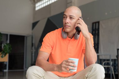 Kafe masasında kahve fincanı ile turuncu polo tişörtlü, kulaklık takan ve akıllı telefon kullanan orta yaşlı yakışıklı kel bir adam. Teknoloji ve yaşam tarzı kavramı.