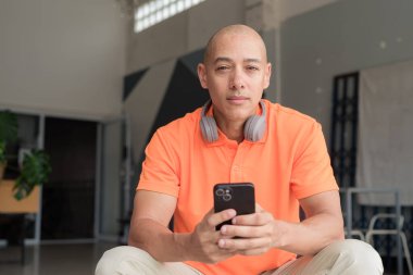 Kafede turuncu polo tişört giyerken cep telefonuyla mesajlaşan kel bir adam.