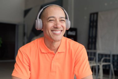 Orta yaşlı yakışıklı, turuncu polo tişörtlü kel bir İtalyan kafe masasında oturmuş kulaklık takıyor ve müzik dinliyor. Günlük yaşam tarzı kavramı.