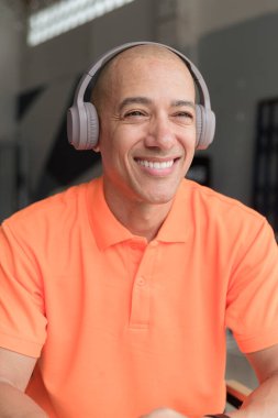 Orta yaşlı yakışıklı, turuncu polo tişörtlü kel bir İtalyan kafe masasında oturmuş kulaklık takıyor ve müzik dinliyor. Günlük yaşam tarzı kavramı.