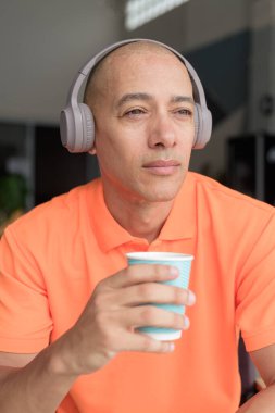 Orta yaşlı yakışıklı, turuncu polo tişörtlü, kahve masasında oturan, kulaklık takan, müzik dinleyen ve elinde kahve fincanı tutan kel bir İtalyan. Günlük yaşam tarzı kavramı.