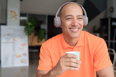 Orta yaşlı yakışıklı, turuncu polo tişörtlü, kahve masasında oturan, kulaklık takan, müzik dinleyen ve elinde kahve fincanı tutan kel bir İtalyan. Günlük yaşam tarzı kavramı.