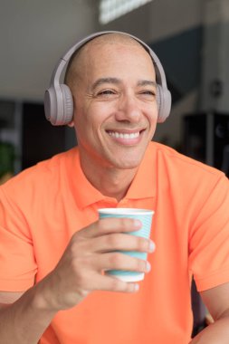 Orta yaşlı yakışıklı, turuncu polo tişörtlü, kahve masasında oturan, kulaklık takan, müzik dinleyen ve elinde kahve fincanı tutan kel bir İtalyan. Günlük yaşam tarzı kavramı.