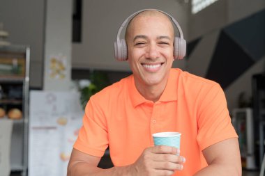 Orta yaşlı yakışıklı, turuncu polo tişörtlü, kahve masasında oturan, kulaklık takan, müzik dinleyen ve elinde kahve fincanı tutan kel bir İtalyan. Günlük yaşam tarzı kavramı.