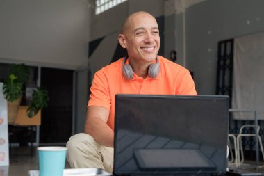 Kafe masasında kahve fincanı takan, kulaklık takan ve dizüstü bilgisayarla çalışan orta yaşlı, kel, turuncu polo tişörtlü yakışıklı bir adam. Uzak çalışma ve teknoloji yaşam tarzı konsepti.