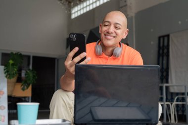 Kafe masasında kahve fincanı ile turuncu polo tişörtlü, kulaklık takan ve dizüstü bilgisayar ve telefonla çalışan orta yaşlı yakışıklı, kel bir adam. Uzak çalışma ve teknoloji yaşam tarzı konsepti.