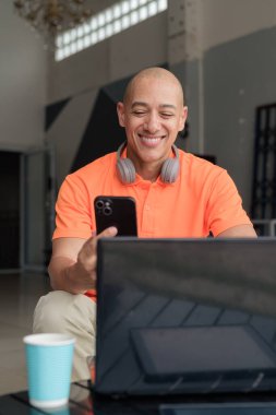 Kafe masasında kahve fincanı ile turuncu polo tişörtlü, kulaklık takan ve dizüstü bilgisayar ve telefonla çalışan orta yaşlı yakışıklı, kel bir adam. Uzak çalışma ve teknoloji yaşam tarzı konsepti.