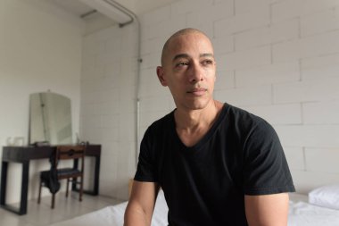 Siyah tişörtlü orta yaşlı, kel bir adam minimalist beyaz bir dairede rahatlarken yatakta oturuyor. Modern bir ev ortamında sıradan bir yaşam tarzı..