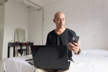 Orta yaşlı, kel, siyah tişörtlü bir adam minimalist beyaz bir dairede dizüstü bilgisayar ve telefon kullanırken yatakta oturuyor. Modern ev işi yaşam tarzı.