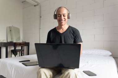 Siyah tişört giyen orta yaşlı, kel bir adam minimalist beyaz bir dairede dizüstü bilgisayar kullanırken yatakta oturuyor. Modern ev işi yaşam tarzı.