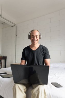 Siyah tişört giyen orta yaşlı, kel bir adam minimalist beyaz bir dairede dizüstü bilgisayar kullanırken yatakta oturuyor. Modern ev işi yaşam tarzı.