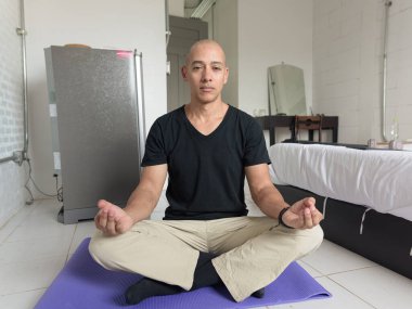 Orta yaşlı, kel, siyah tişörtlü bir adam yoga ve meditasyon yaparken yoga minderinde yan yana oturuyor. Ev ortamında rahatlama ve farkındalık yaşam tarzı