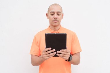 Portakal rengi polo tişörtlü yakışıklı, orta yaşlı, kel bir adamın dijital tablet bilgisayar kullanarak beyaz duvar arkaplanının stüdyo portresi. Günlük yaşam tarzı ve moda konsepti.