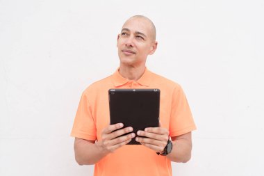 Portakal rengi polo tişörtlü yakışıklı, orta yaşlı, kel bir adamın dijital tablet bilgisayar kullanarak beyaz duvar arkaplanının stüdyo portresi. Günlük yaşam tarzı ve moda konsepti.