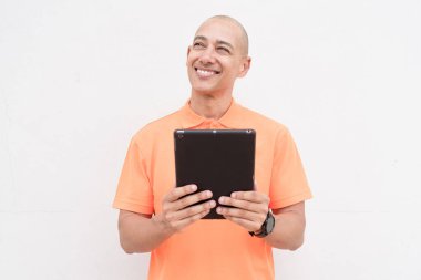 Portakal rengi polo tişörtlü yakışıklı, orta yaşlı, kel bir adamın dijital tablet bilgisayar kullanarak beyaz duvar arkaplanının stüdyo portresi. Günlük yaşam tarzı ve moda konsepti.